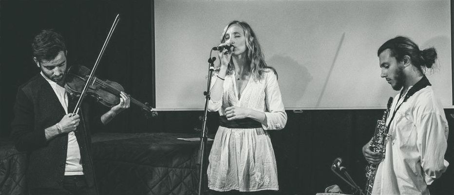 Will on violin, Sylvia singing at the microphone, Seb holding a soprano saxophone, on a dim stage